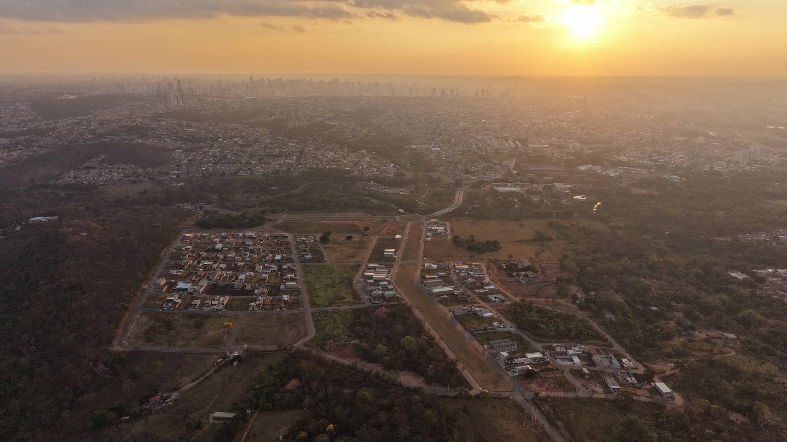 Vista aérea do Village Campos Verdes ao entardecer: quadras com ruas asfaltadas e casas construídas no primeiro plano, áreas ainda vazias em volta e o horizonte de Goiânia ao fundo.