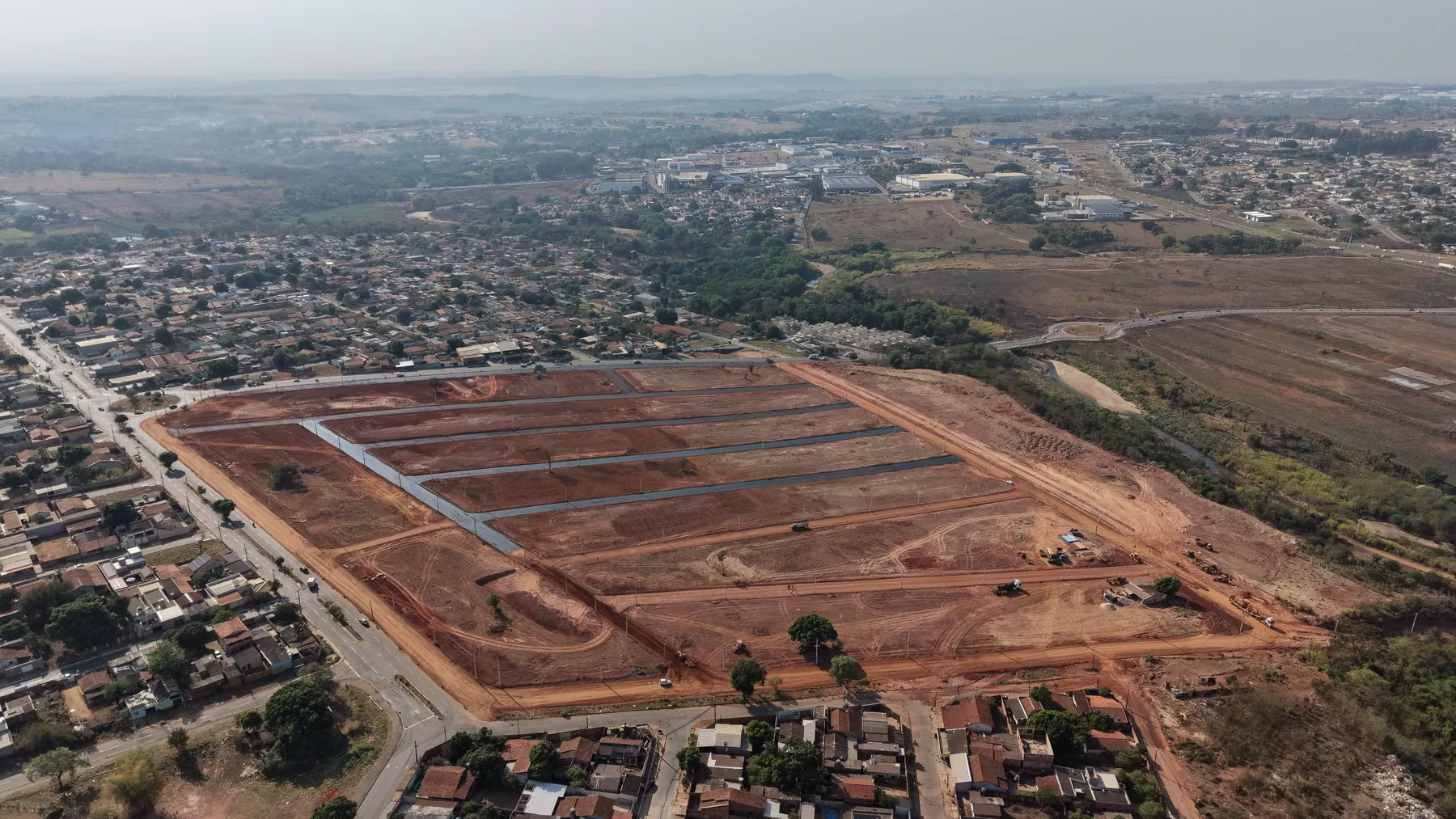 Vista aérea do Residencial Boulevard em 1º de setembro de 2026: a área loteada em terra vermelha, dividida em quadras compridas por ruas já abertas, com postes ao longo delas; em volta, um bairro construído, e ao fundo a cidade.